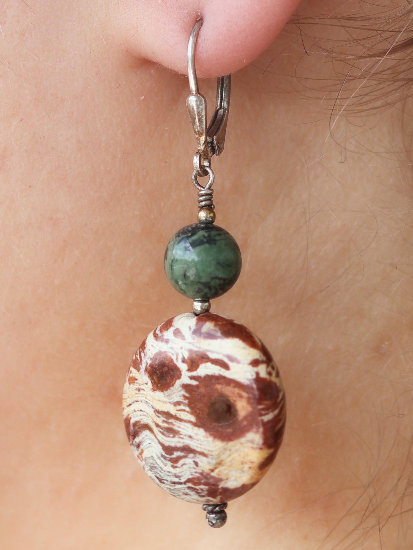 Close-up of an earring with a rust and cream colored, marbled, round and flat bead with a small, round, green bead