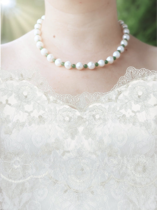 Pearl necklace with each pearl separated by small, round emerald beads - worn by a person in a white lace wedding dress with a blurred green background