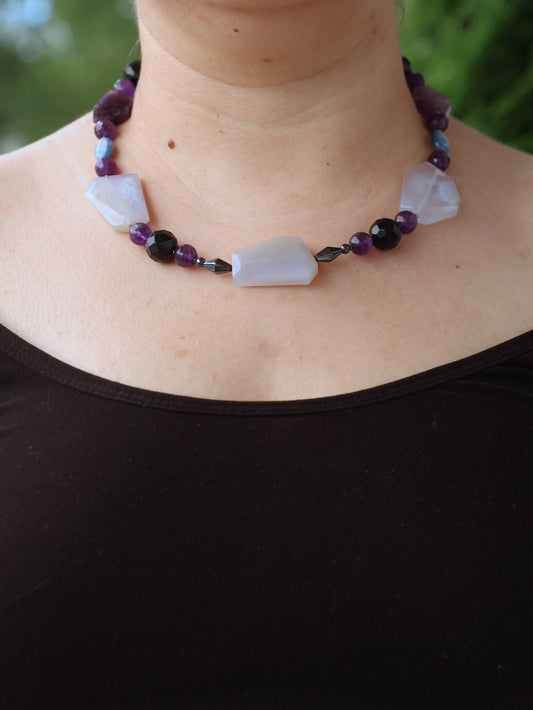 Necklace with deep violet, black, and chunky light blue beads on a person wearing a black top against a blurred green background